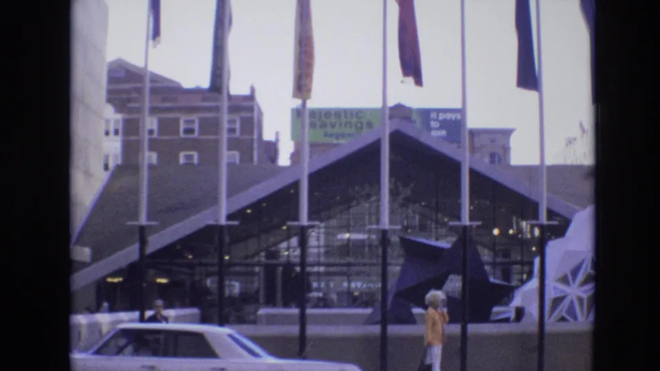 1974 Denver street view with tall international flagpoles, an angular glass-fronted building, background Majestic Savings signage, a parked car, and a woman walking past.