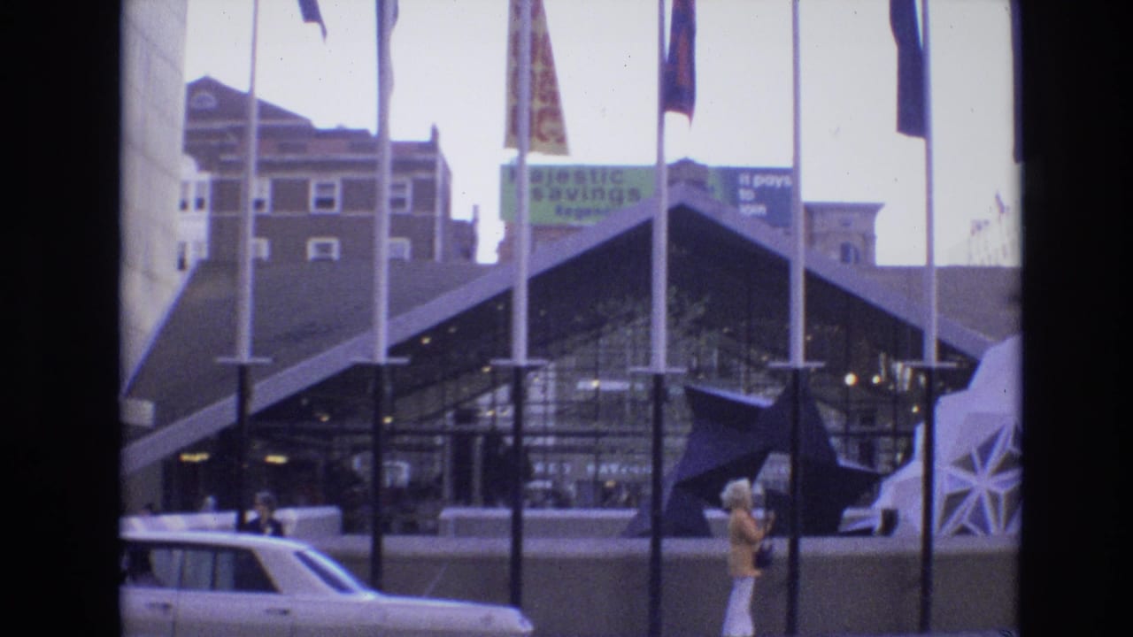 1974 Denver exterior with international flags, a dark geometric glass facade, a visible 1970s car, background signs, and a woman in light trousers walking along the street edge.