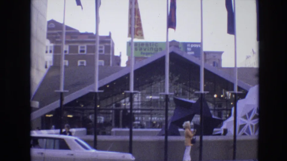 1974 Denver exterior with international flags, a dark geometric glass facade, a visible 1970s car, background signs, and a woman in light trousers walking along the street edge.
