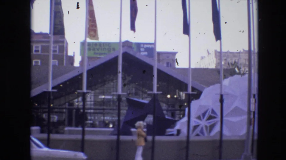 1974 Denver frame showing flagpoles in front of an angular building, a white geodesic dome-like structure at right, a woman walking, an older car, and background commercial signage.