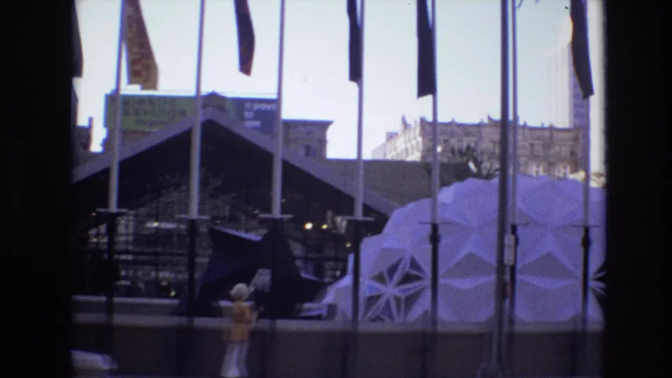 1974 Denver street view with tall flags, dark modern glass architecture, a white dome-like geometric structure, a woman in period clothing, and background urban buildings.
