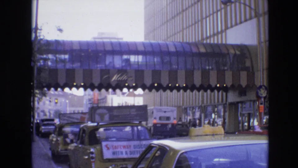 1974 Denver downtown traffic scene with yellow taxis and classic cars below a glass-enclosed pedestrian bridge, with pedestrians and storefront signage in the distance.