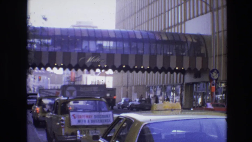 1974 Denver street frame with a yellow taxi carrying a roof sign, other classic cars, pedestrians, and a glass pedestrian bridge spanning the roadway.