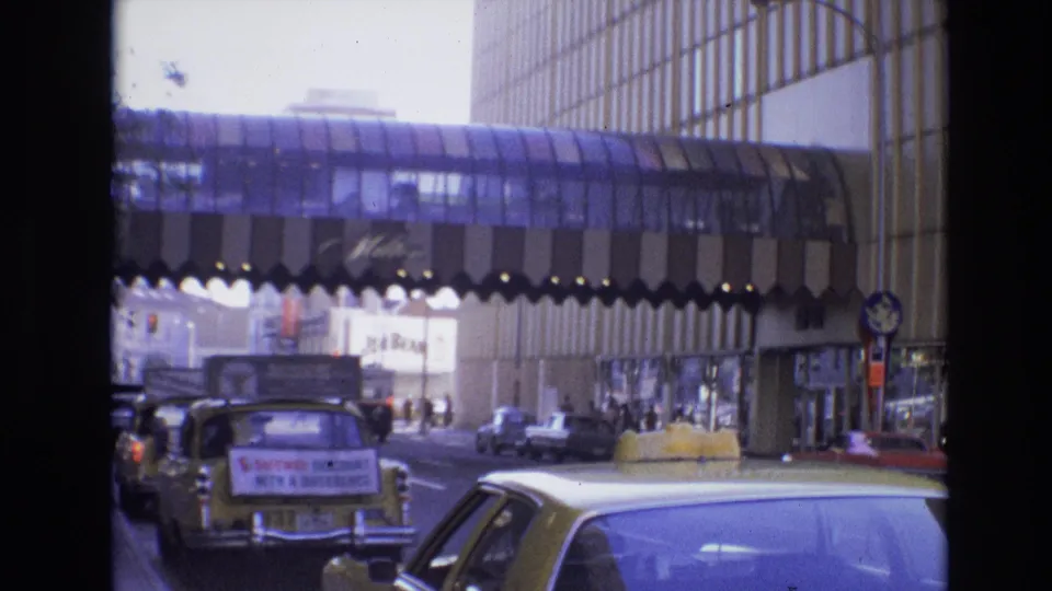 1974 Denver home-movie frame showing a yellow taxi and other classic cars in traffic beneath a glass pedestrian bridge, with pedestrians and commercial signage at street level.