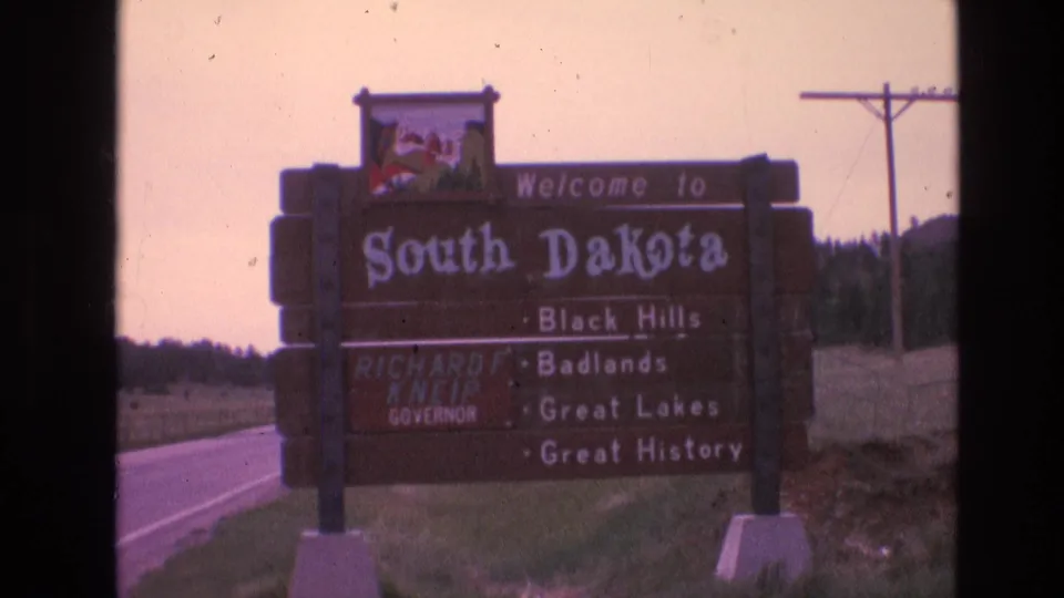 1974 8mm frame of a South Dakota welcome sign beside a rural highway, with Black Hills, Badlands, Great Lakes, and Great History listed on the board.