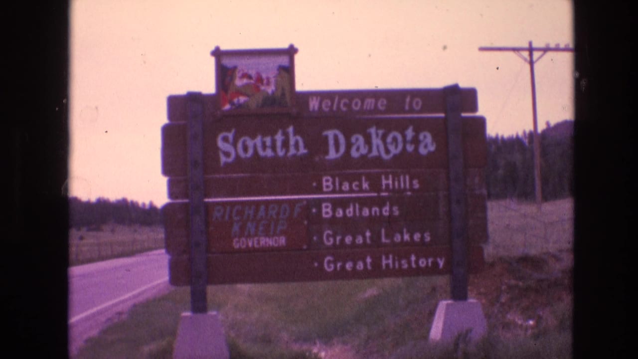 The South Dakota welcome sign appears from a moving roadside viewpoint, with Richard F. Kneip named on the board and power lines visible behind it.