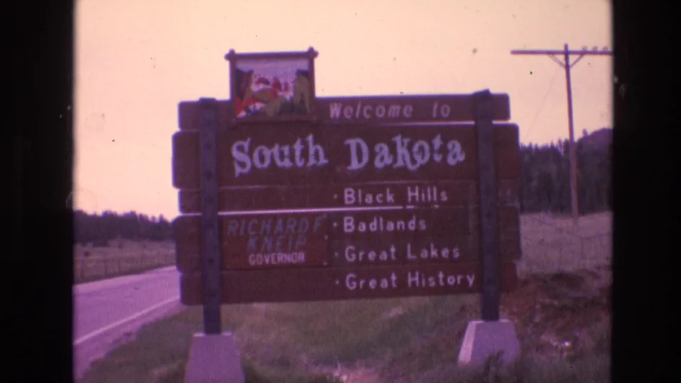 The South Dakota welcome sign appears from a moving roadside viewpoint, with Richard F. Kneip named on the board and power lines visible behind it.