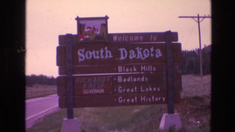 A closer 8mm view of the South Dakota highway welcome sign shows the large state name, attraction list, grassy shoulder, and road edge.