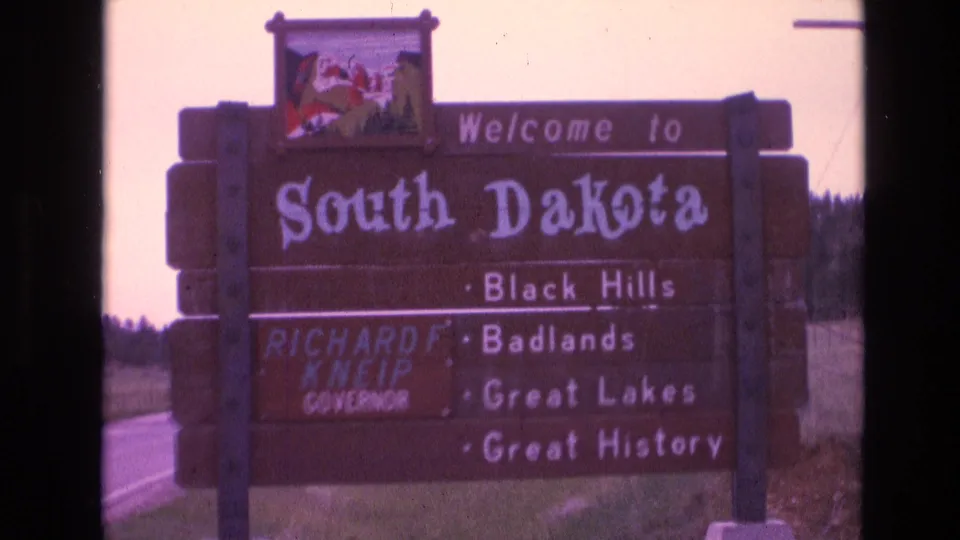 The South Dakota welcome sign fills much of the frame, with weathered wooden posts, attraction text, and a rural roadside background visible.