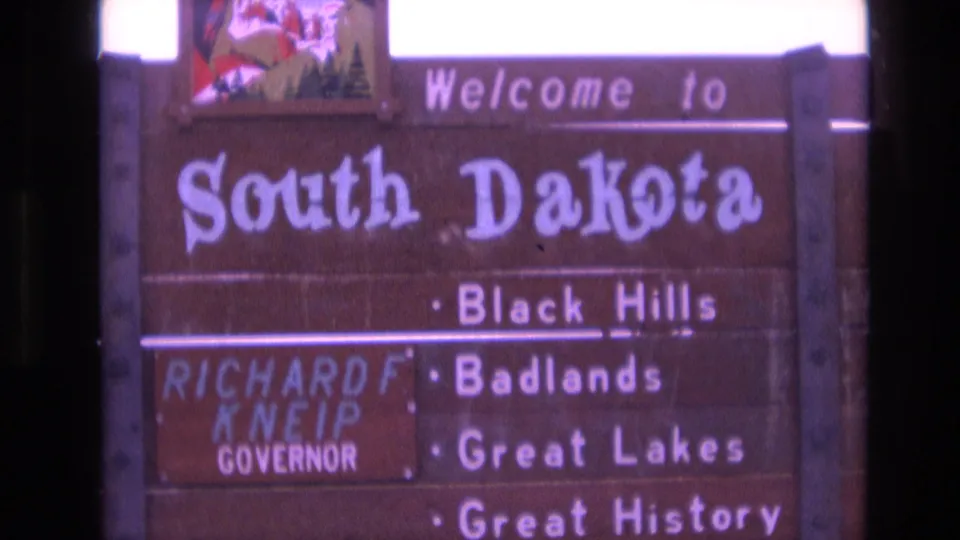 Close 8mm frame of the South Dakota welcome sign in 1974, with the state name dominating the frame and the Richard F. Kneip governor panel visible at left.