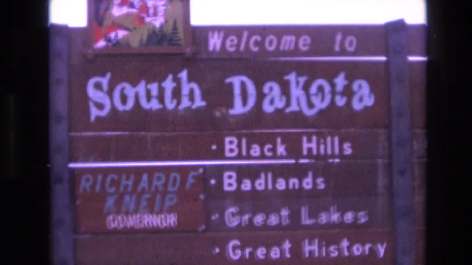 Tight final frame of the 1974 South Dakota welcome sign, with white attraction text and the governor name panel partially softened by focus and motion.