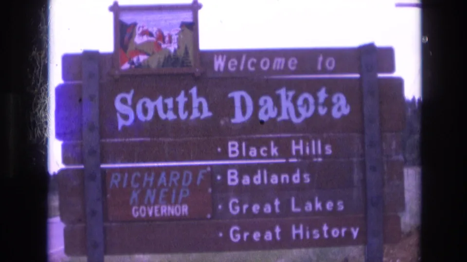1974 8mm frame showing the full wooden South Dakota welcome sign with scenic emblem, Governor Kneip panel, roadside edge, grass, and bright overexposed sky.