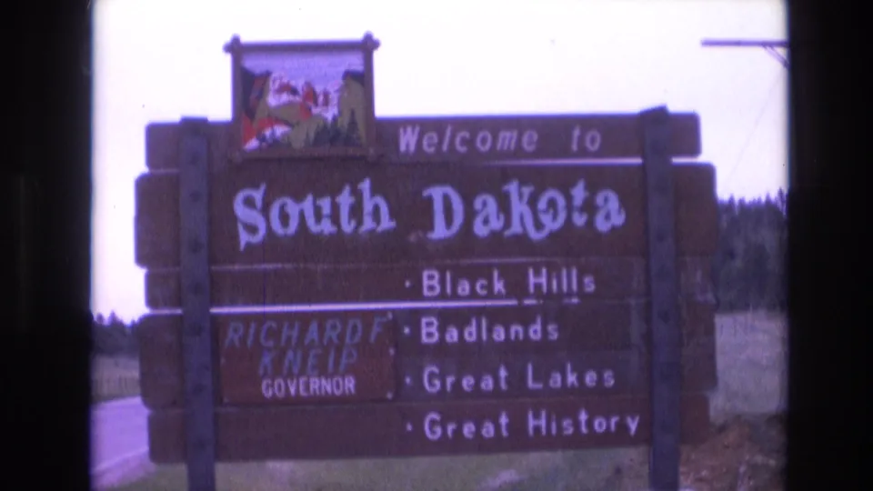 1974 8mm frame of the South Dakota welcome sign from a slightly wider roadside angle, with the Governor Kneip panel, attraction list, grass verge, and sky visible.