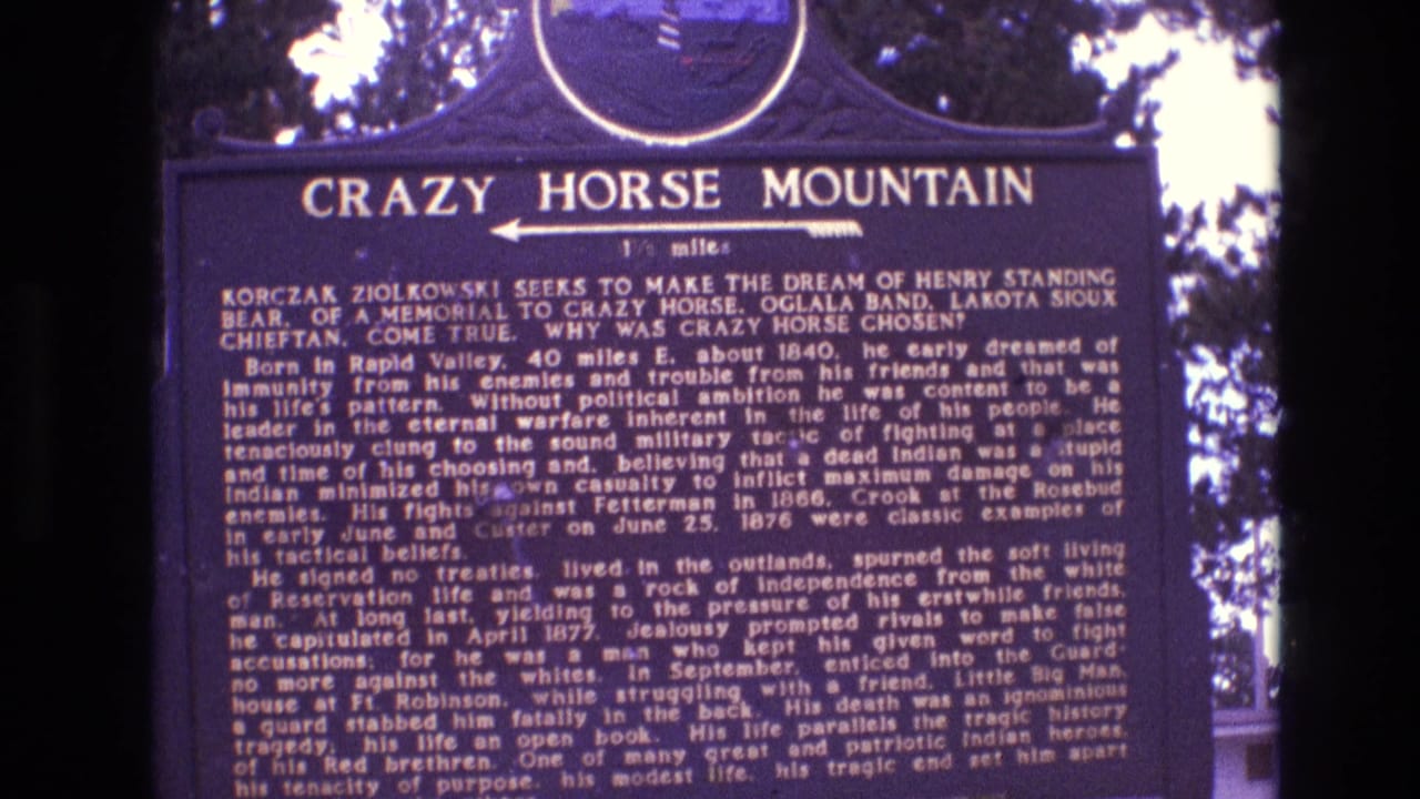 1974 South Dakota 8mm frame showing a dark blue Crazy Horse Mountain historical marker, with tree branches and bright sky behind the sign.
