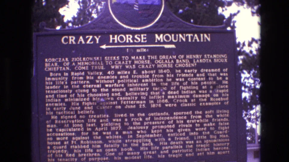 1974 South Dakota 8mm frame showing a dark blue Crazy Horse Mountain historical marker, with tree branches and bright sky behind the sign.