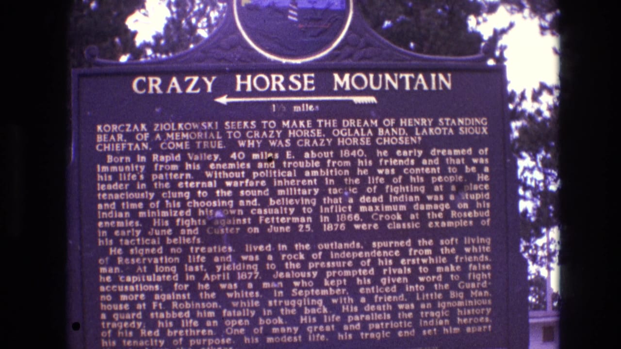 Tightly framed 1974 8mm view of the Crazy Horse Mountain plaque in South Dakota, showing white lettering on a dark sign with foliage in the background.