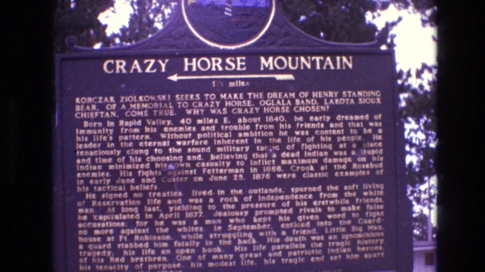 Handheld 8mm frame from the South Dakota marker, with the Crazy Horse Mountain title, arrow, and historical text visible through soft focus and color shift.