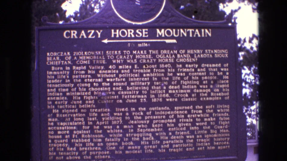 Final 1974 8mm frame holding on the Crazy Horse Mountain historical marker in South Dakota, with dense plaque text and surrounding tree silhouettes.