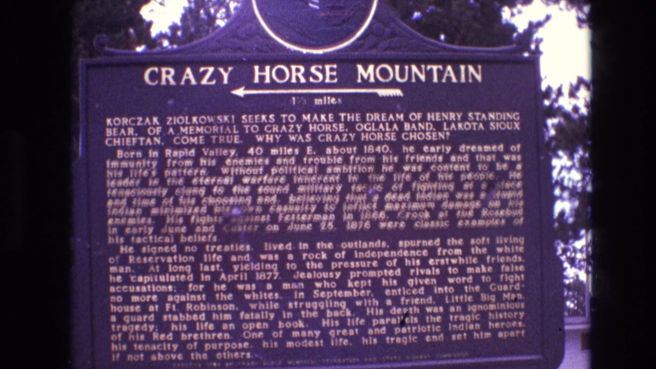 1974 South Dakota 8mm frame showing a dark blue Crazy Horse Mountain marker with dense white interpretive text, a left-pointing arrow, and trees behind it.