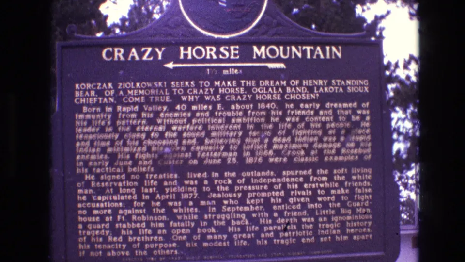 1974 South Dakota 8mm frame showing a dark blue Crazy Horse Mountain marker with dense white interpretive text, a left-pointing arrow, and trees behind it.