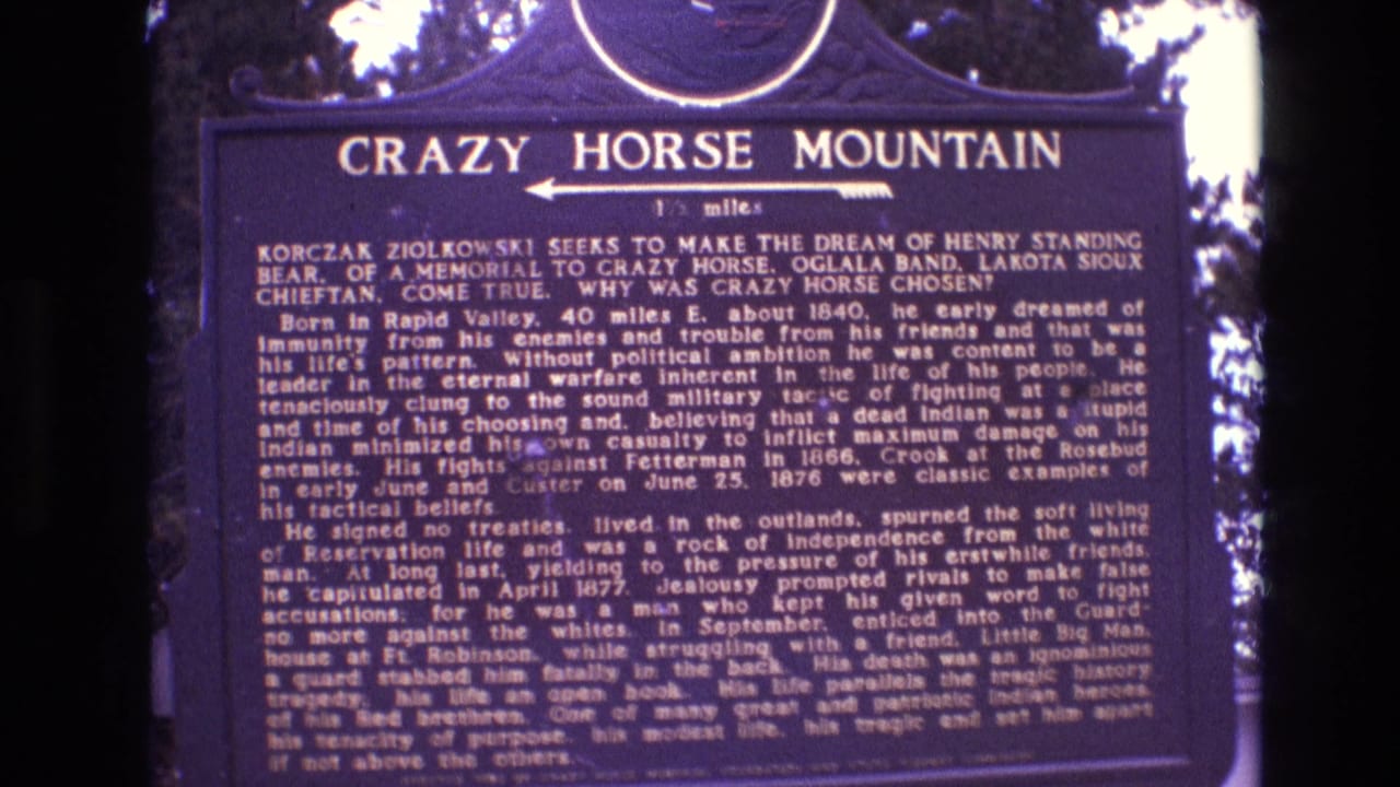 1974 South Dakota 8mm view of the Crazy Horse Mountain historical plaque, with white lettering on a dark blue sign and wooded background visible at the edges.