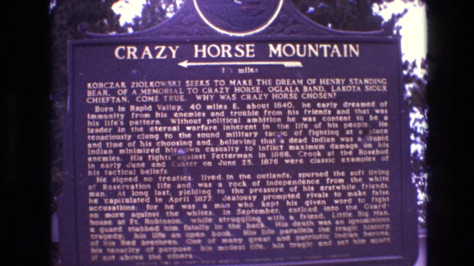 1974 South Dakota 8mm view of the Crazy Horse Mountain historical plaque, with white lettering on a dark blue sign and wooded background visible at the edges.