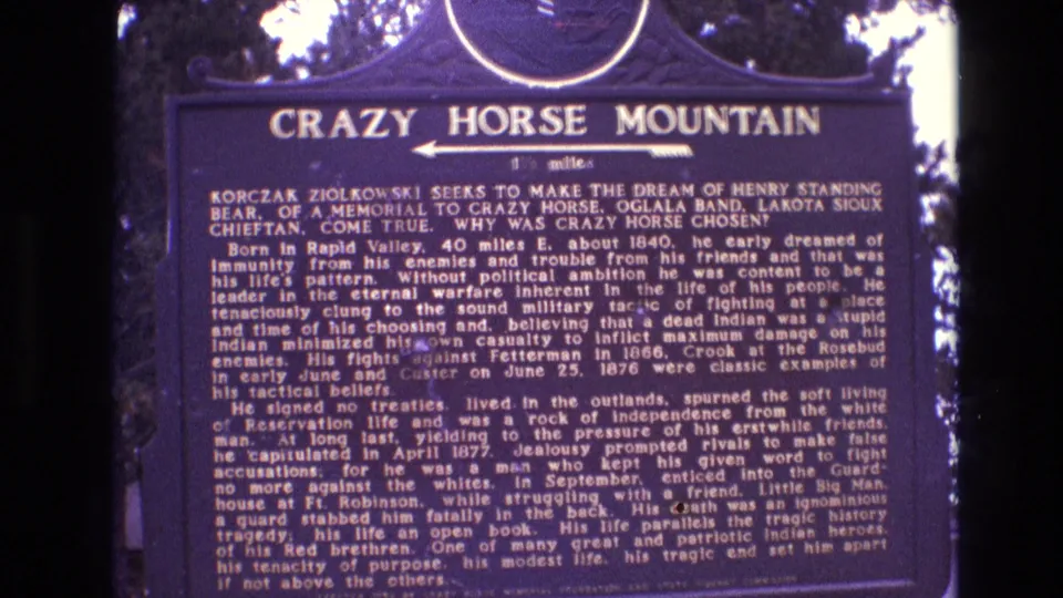 1974 South Dakota home-movie frame holding on the Crazy Horse Mountain marker, emphasizing the title, arrow, and compact historical text on the sign face.