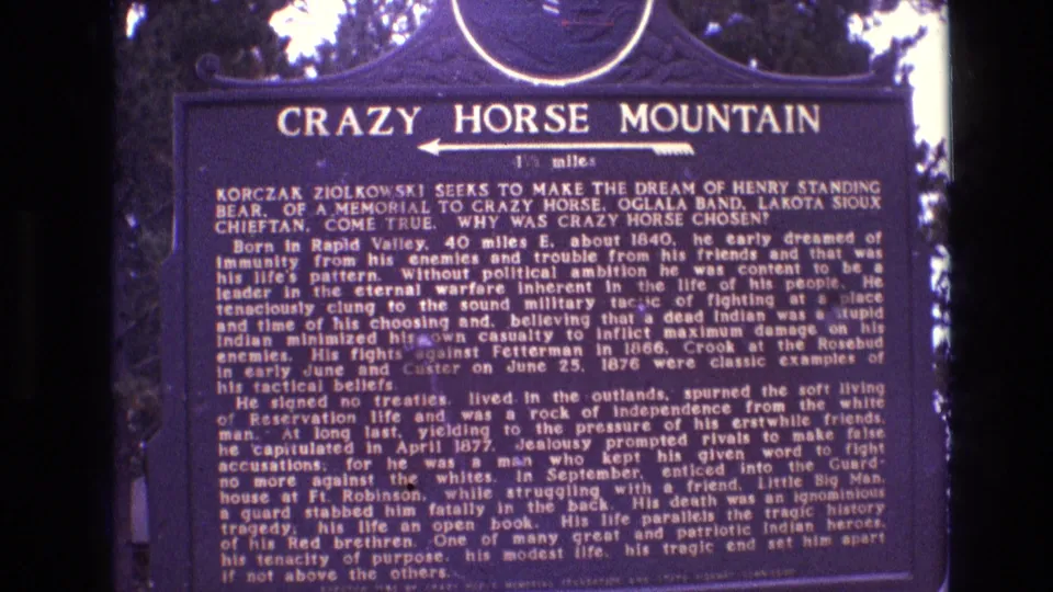 Final 1974 South Dakota 8mm frame of the Crazy Horse Mountain historical marker, showing the dark blue plaque, white text, arrow, and surrounding trees in soft home-movie focus.