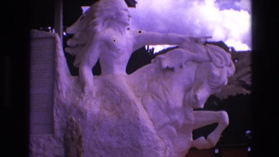 1974 South Dakota home-movie frame showing the outdoor rider-and-horse sculpture with strong purple color cast, rough stone base, and cloudy sky.