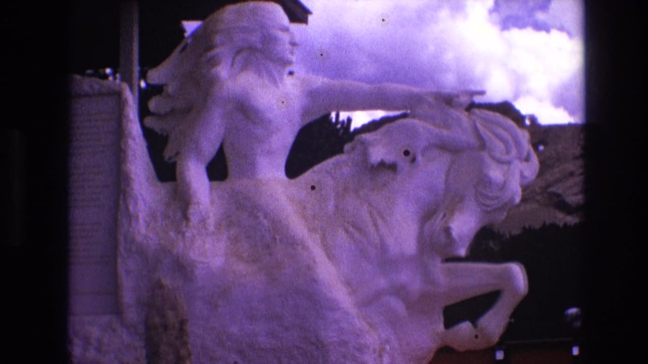1974 South Dakota 8mm frame showing a pale stone sculpture of a Native American figure riding a rearing horse, seen from a visitor angle under a purple-tinted cloudy sky.