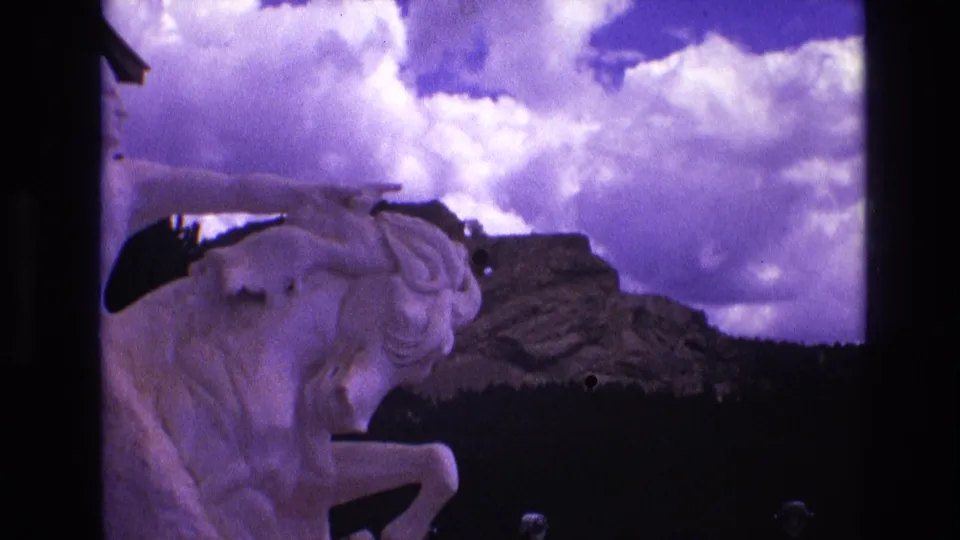 The final frame holds on the white equestrian statue in South Dakota, with dark frame edges, a rocky outcropping, and a cloud-heavy purple-blue sky.