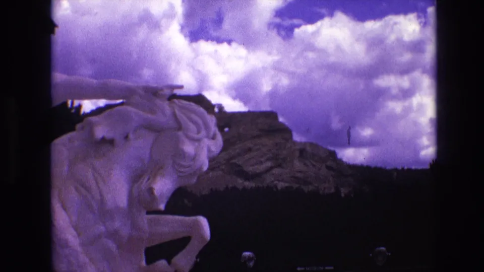 1974 South Dakota 8mm frame showing the white horse statue near the left edge, a layered rocky hillside, a dark tree band, and billowing clouds.