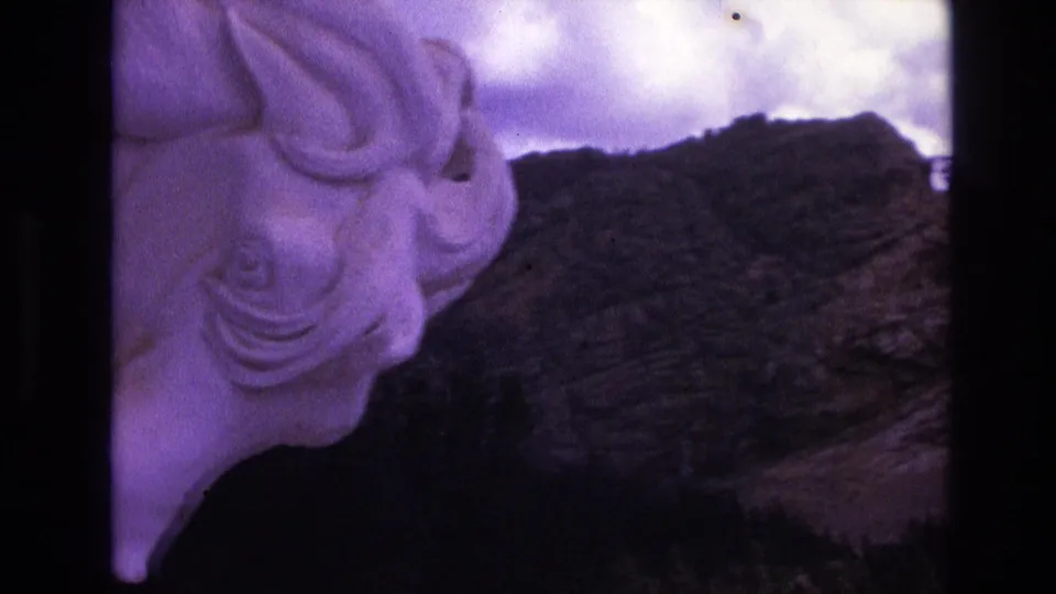 1974 South Dakota home-movie frame moving closer to the white horse statue, with its sculpted head and neck against a rocky hillside and pale clouds.