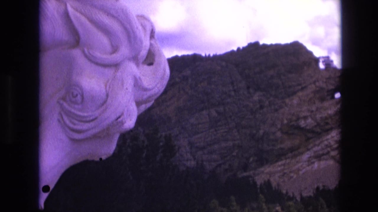 1974 South Dakota 8mm frame showing a pale carved stone face along the left edge with a rugged mountain ridge, evergreen trees, and purple-shifted sky in the background.