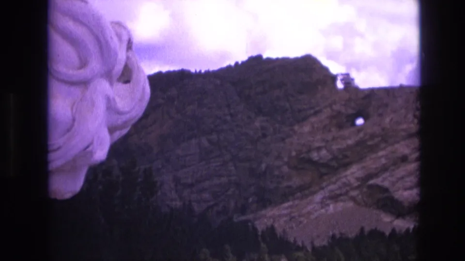 1974 South Dakota 8mm frame with the carved face partly cropped at left and a rugged rock formation with a small circular opening under a cloudy purple-tinted sky.