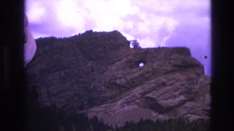 1974 South Dakota 8mm frame as the camera pans away from the carved face toward a layered mountain ridge, evergreen trees, and a distinct small hole in the rock.