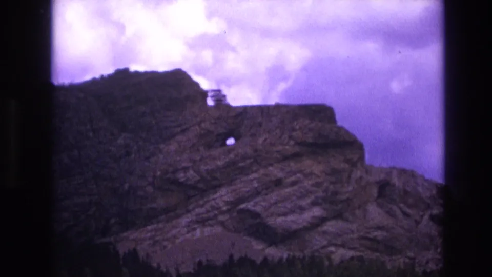 1974 South Dakota 8mm frame focused on the rugged rock formation, showing layered stone, a small circular opening, a dark tree line, and purple-tinted clouds.