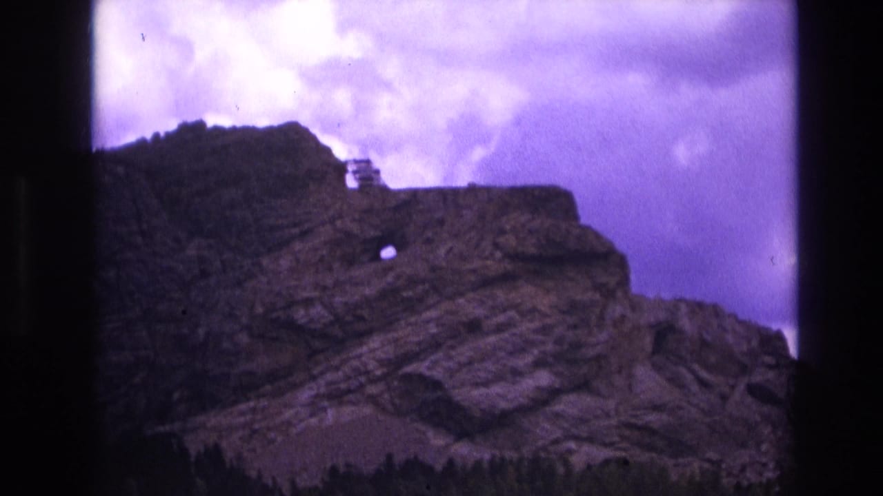 1974 South Dakota 8mm frame showing a rugged layered rock formation, a small circular opening in the cliff face, dark trees at the base, and a purple-tinted cloudy sky.