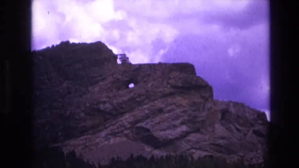 1974 South Dakota 8mm frame showing a rugged layered rock formation, a small circular opening in the cliff face, dark trees at the base, and a purple-tinted cloudy sky.