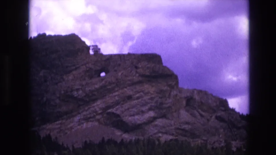 1974 South Dakota 8mm frame of the same cliff from a slightly shifted view, with the round rock opening, stratified stone layers, summit detail, forest edge, and lavender cloud cover visible.