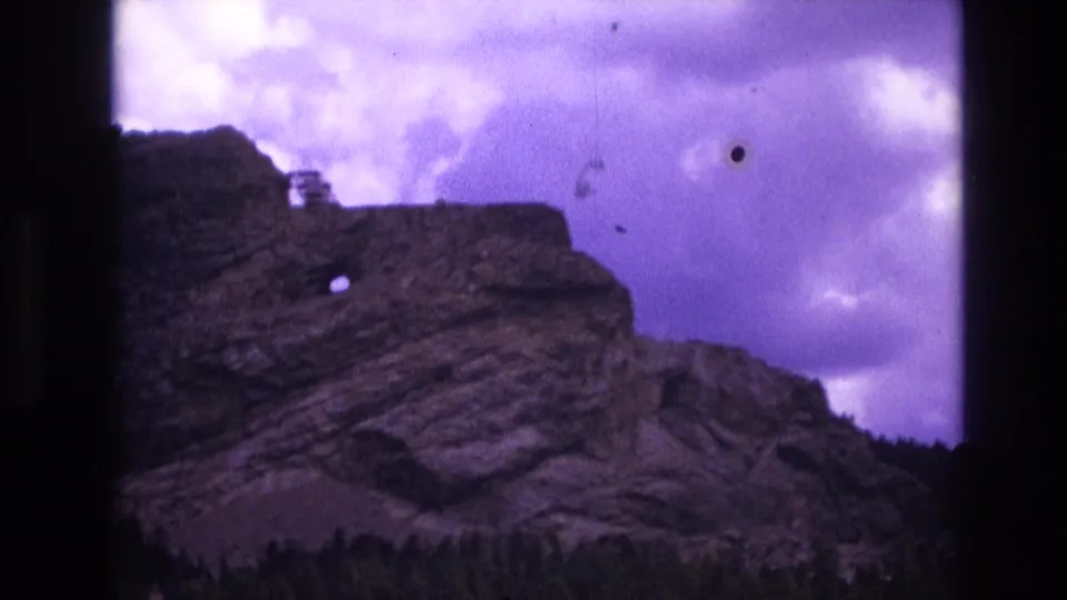 1974 South Dakota 8mm frame with a close tourist view of the layered rock formation, visible circular hole, dark vegetation along the bottom edge, dust specks, and purple overcast sky.