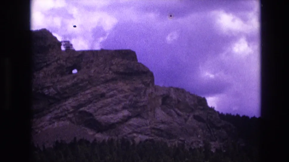 1974 South Dakota 8mm frame holding on the rugged cliff, round rock opening, small ridge-top structure, dark tree line, and purple-tinted cloud cover within visible film-frame borders.