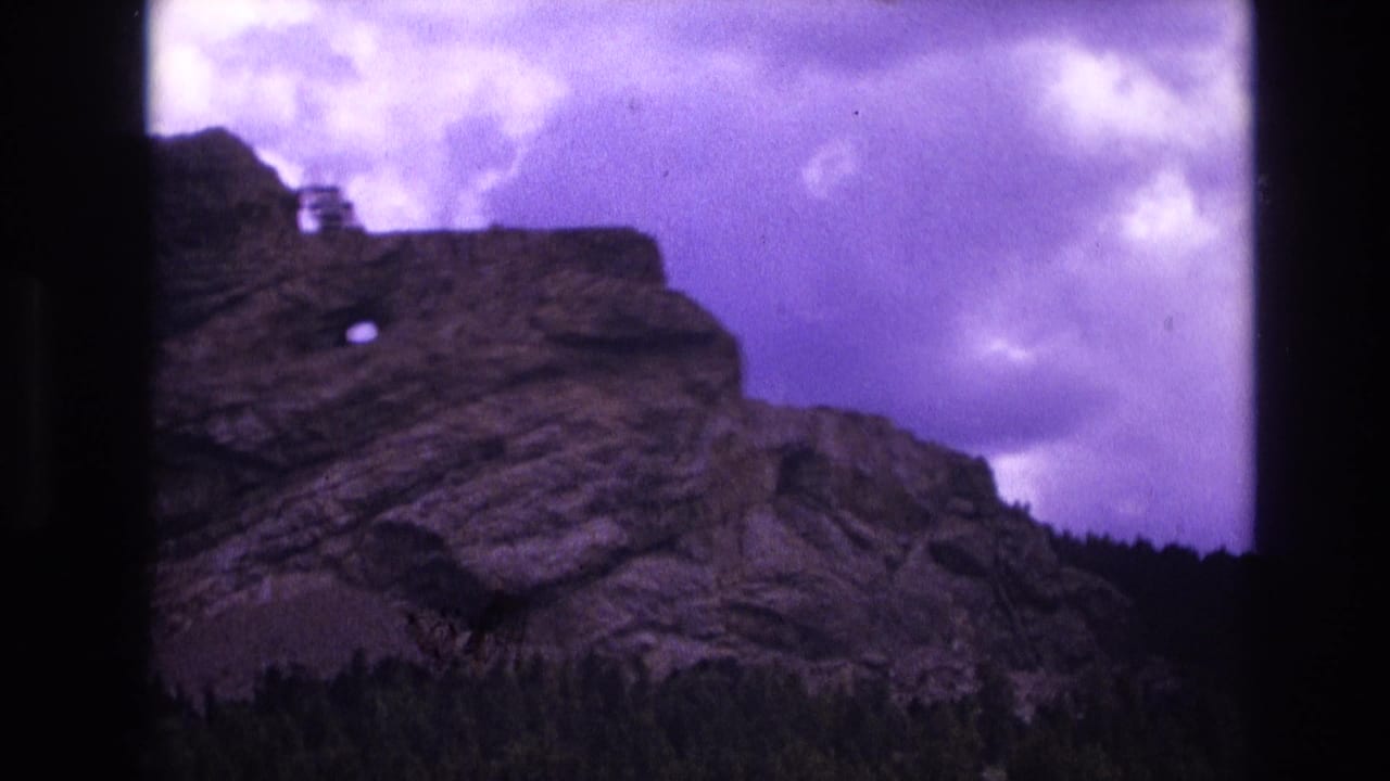 1974 8mm South Dakota home-movie frame showing a stratified rocky outcropping rising above evergreen forest beneath a purple-tinged cloudy sky.