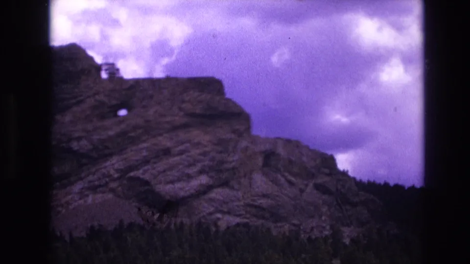 1974 8mm South Dakota home-movie frame showing a stratified rocky outcropping rising above evergreen forest beneath a purple-tinged cloudy sky.