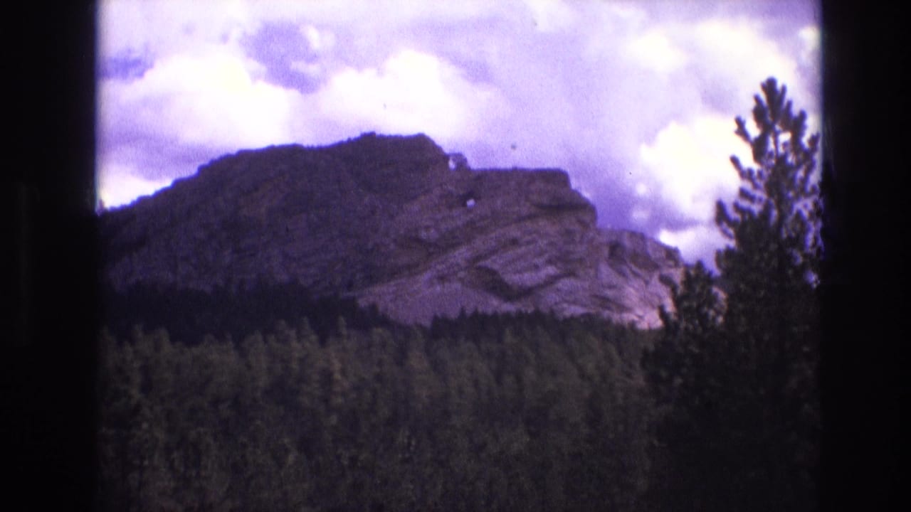 1974 South Dakota 8mm frame with a broad rock face above dense forest, cloudy sky overhead, and a tall conifer along the right side of the composition.