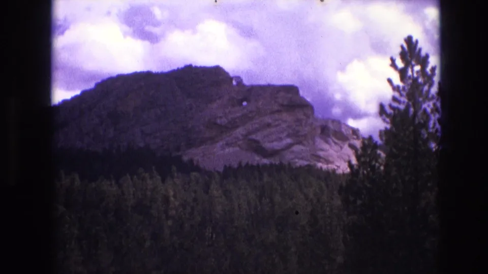 South Dakota 1974 home-movie frame showing layered rock above a wooded slope, with a tall tree at right and strong magenta-purple film color shift.
