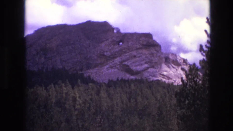 Final 1974 8mm South Dakota frame holding on the rocky outcropping above evergreen forest, with cloudy sky, visible film borders, and a small pale feature near the ridge.