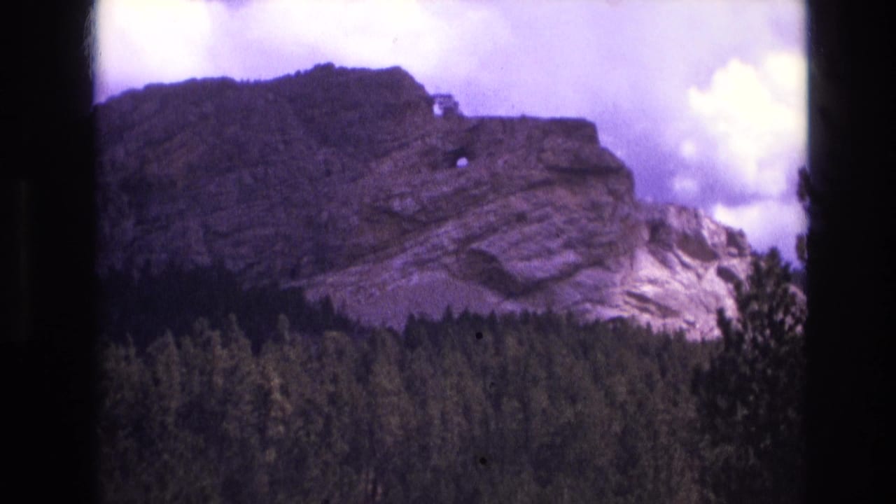 1974 8mm South Dakota home-movie frame showing a layered rock face above a dark evergreen slope, with purple color shift and visible film-edge shadowing.