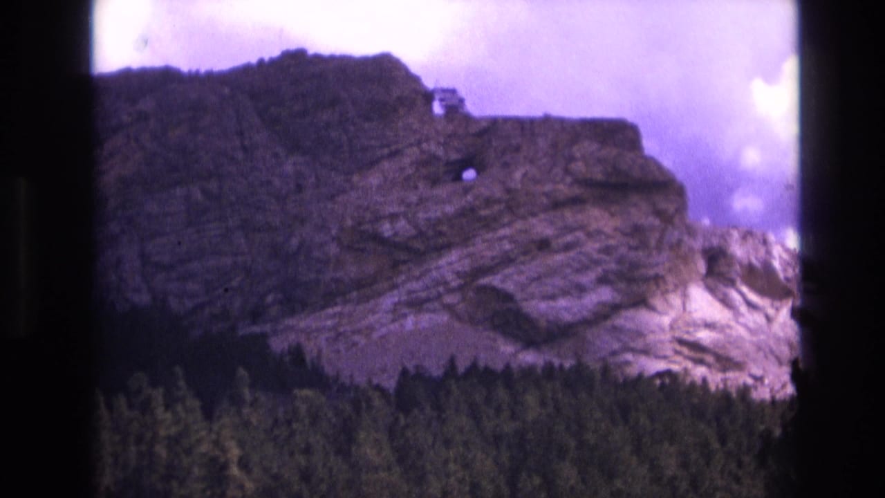Rocky mountain face with evergreen forest below and clouds behind it.