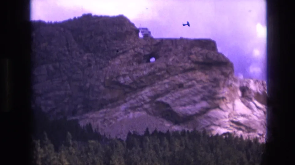 1974 8mm South Dakota home-movie frame showing a closer view of the layered cliff, evergreen trees along the bottom edge, a small vehicle-like shape near the ridge, and pronounced film grain.